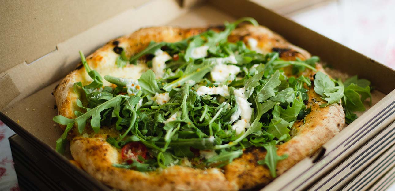 A pizza with arugula on in in a pizza box on a table.